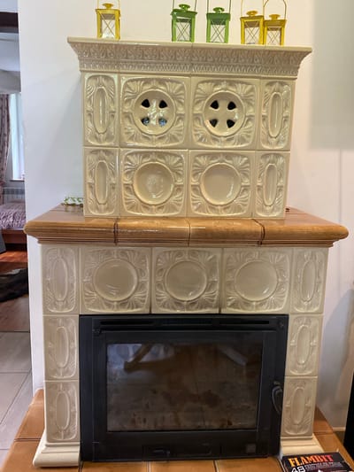 The image shows a fireplace with decorative ceramic tiles, located in Biała Wieś, Poland. The fireplace has a wooden mantel and a black metal insert with a glass door. The ceramic tiles are cream-colored and feature embossed floral and geometric patterns. On top of the fireplace, there are several small decorative lanterns. The background suggests an indoor setting, possibly a residential interior, with part of a wall and a window visible on the left. A box of fire starters labeled "FLAMBIT" is placed on the wooden surface to the right of the fireplace. The lighting is natural, coming from the window and ambient room light.