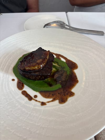 A close-up, overhead shot shows a plate of food in what appears to be a restaurant in Carboneras, Spain. The dish features a piece of meat, possibly steak, topped with a slice of seared foie gras, all coated in a dark glaze. The meat rests on a bed of vibrant green puree, likely vegetables. A brown sauce is artfully drizzled around the puree. The white, textured plate has a subtle circular pattern. In the background, a blurred spoon rests on another white plate. Partially visible figures in the background suggest other diners or staff in the restaurant. The overall scene is a well-plated gourmet dish presented in a sophisticated dining environment.