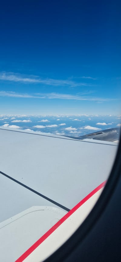 The image is an aerial view from an airplane window, looking out at the sky and clouds. The airplane's wing is prominently visible in the foreground, with a white surface and a red stripe along its edge. The sky is a deep blue, with scattered white clouds forming a layer above the horizon. The environment appears to be clear and sunny, with good visibility. There are no people or specific landmarks identifiable that would indicate the location in Groß-Zimmern, Germany. The image captures a serene moment during an airplane journey.