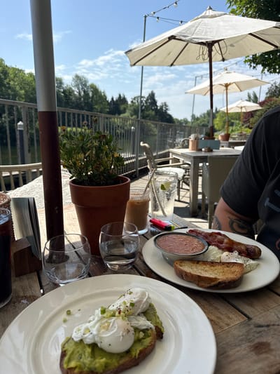 This image shows a close-up view of a breakfast spread on a wooden table, with an outdoor setting in the background. The subjects are two plates of food, drinks, and a potted plant. One plate features avocado toast topped with poached eggs and green onions, while the other has a full English breakfast with bacon, sausage, baked beans, a piece of toast, and poached eggs. A glass of iced water with lime, another drink with a straw, and two empty wine glasses are also on the table. The potted plant sits in a terracotta pot.

In the background, several white patio umbrellas are visible, suggesting a restaurant or cafe with outdoor seating. Beyond the umbrellas, there is a railing and lush green trees under a bright blue sky with scattered clouds, indicating it is daytime and likely pleasant weather. The overall scene depicts a meal being enjoyed outdoors.

No visible text is present in the image. The orientation of the image is such that the sky and umbrellas appear on the left, and the food is on the right. The image appears to be taken from the perspective of someone sitting at the table. The location context is Maidenhead, United Kingdom, which aligns with the presence of outdoor dining and typical greenery.