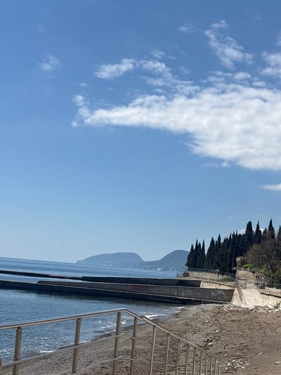 The image depicts a beach scene in Semidvorye, Ukraine, under a partly cloudy sky. The foreground includes a metal railing and part of a concrete structure extending into the water. Water meets the concrete structure, with waves visible. Beyond the water, there is a distant mountain. Trees line the top edge of the concrete structure. The sky is blue with white clouds.