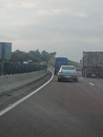 A highway scene presents several vehicles on the road. A silver car is closely following a blue truck branded with "Locamed." Ahead, a larger, darker truck is partially visible. The road is asphalt with a white line marking the edge. Beyond the barrier, trees dot the landscape under a cloudy sky. The image appears to have been taken from another vehicle on the same road.