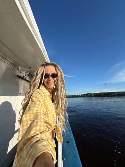 A woman with long blonde dreadlocks and dark sunglasses smiles directly at the camera, taking a selfie on a boat in the city of Dumino, Russia. She wears a yellow and white checkered button-down shirt, tied at the waist, over a white top. Her left side is against a white boat wall, which displays a red "П-2" marking and casts sharp shadows. To her right, a blue railing borders a wide expanse of dark blue water with ripples and foam. The water extends to a distant, tree-lined shore, above which a tall, slender lattice communication tower is visible. The sky above is clear blue with a few wispy clouds and two birds in flight, one appearing to be a seagull on the right side of the frame.