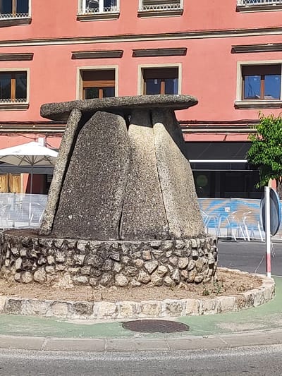 This is a medium shot, eye-level photograph taken outdoors during the day. The focal point is a large, stone monument that resembles a dolmen. The monument is made of rough, grey stones, with a large flat slab on top supported by three large, triangular upright stones. The base of the monument is encircled by a circular wall constructed from smaller, irregular stones. The monument is situated in what appears to be a roundabout or public square, with a paved road visible in the foreground and a light green painted area surrounding the monument.
In the background, a building with a pink facade is visible. The building has several windows with dark wooden frames and white ornate grilles. Below the monument, a white umbrella from an outdoor seating area and some metal fencing are partially visible. To the right of the monument, a small green tree and a white pole with a round mirror are present, along with some fencing with a blue and orange pattern.
The lighting suggests it is a sunny day. The overall mood is one of a historic or cultural landmark in a public space. There is no visible text in the image. The location context of Valencia de Alcántara, Spain, suggests this monument is likely a historical or archaeological representation.