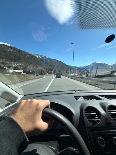 The image depicts a first-person view from the driver's seat of a vehicle traveling on a road in Saint-Chaffrey, France, under clear weather conditions. A right hand, featuring visible arm hair and clad in a dark sleeve, grips the black steering wheel. Portions of the vehicle's black dashboard are visible, including air vents, a red hazard light button, a control panel, and an open storage compartment containing papers. The windshield shows reflections, including a faint outline of a hand, and a black circular object attached to its upper right corner.

Ahead, a two-lane asphalt road with white lane markings extends forward. A dark-colored SUV or minivan is visible in the same lane. To the left, the roadside transitions to a grassy area with multiple streetlights, road signs, and several residential or commercial buildings nestled at the base of tree-covered mountains. A distinctive white arched structure, characteristic of the entrance to the Serre Chevalier valley in Saint-Chaffrey, is visible spanning the road in the mid-distance. To the right, a metal guardrail runs parallel to the road. The background is dominated by towering mountains, partially covered in snow on their upper slopes and dense evergreen forests lower down, all beneath a bright blue sky with scattered white clouds.