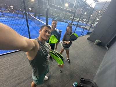 The scene depicts an indoor padel tennis facility identified as Padel ONE Mülheim, located at Mintarder Str. 270, 45481 Mülheim an der Ruhr, Germany. The environment features blue padel courts with white lines, black perimeter fencing, and black structural walls. The flooring in the immediate foreground is grey carpet.

Three individuals are posing for a photograph, likely a selfie.
1.  In the foreground, a male with dark hair and light olive skin, wearing a grey sleeveless Under Armour top and light green shorts, holds a neon green and black padel racket and the camera. He is smiling towards the lens.
2.  To his left, a second male with dark hair and light olive skin, wearing a dark green Adidas t-shirt and dark shorts, holds a neon green and black padel racket. He is looking towards the camera with a slight smile.
3.  To the right, a female with dark hair tied back in a ponytail and light olive skin, wearing an olive-brown oversized t-shirt and light grey Adidas shorts, holds a neon green and black padel racket. She is also looking towards the camera with a slight smile.

All three subjects appear to be young adults. In the background, additional padel courts are visible, displaying "CUPRA" and "Padel ONE" branding on banners. In the immediate foreground on the right, a black bag and a plastic water bottle are placed on a dark surface. A green cylindrical bin is positioned against the black wall. The facility is illuminated by overhead artificial lighting.