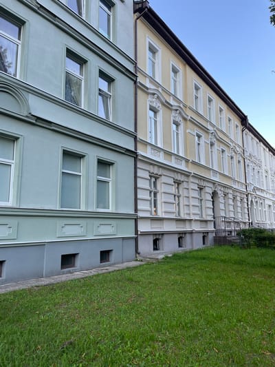The image shows a side view of a row of connected apartment buildings in Sovetsk, Russia, under a clear blue sky. The buildings are constructed with distinct architectural styles and colors. The closest building on the left has a pale green facade with grey detailing near the bottom and around the windows. It features multiple windows, some with blinds, and decorative architectural elements. Adjacent to it, and extending into the distance, are two buildings with creamy yellow facades and white decorative trim around the windows and along the levels. These buildings exhibit more ornate detailing, including sculpted elements above some of the windows. The ground in front of the buildings is covered with a lush green lawn. The scene appears to be a residential area, captured in what appears to be late afternoon or early evening light.