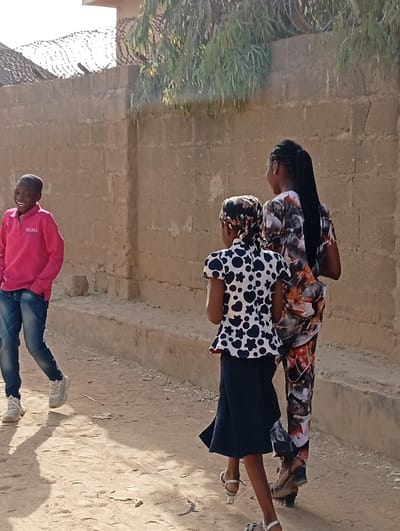 The scene depicts an outdoor setting on a dirt path in Gayawa, Nigeria. A tan concrete block wall, topped with barbed wire and overhung by green foliage, forms the background. The ground is light brown dirt, showing strong shadows indicative of bright daylight.

Three individuals are present. On the left, a young male with dark skin and short hair walks towards the right, smiling broadly with an open mouth. He wears a pink long-sleeved polo shirt, blue jeans, and white athletic shoes, with both hands in his pockets. In the center, a young female with dark skin walks away from the viewer. She wears a patterned headscarf, a white short-sleeved top with dark blue heart and flower patterns, a dark blue pleated skirt, and light-colored open-toed sandals. To her right and slightly behind her, another young female with dark skin also walks away from the viewer. She has long dark braided hair and wears a multi-colored patterned t-shirt and matching patterned pants, along with dark wedge sandals. Both females are in motion, walking in the same direction, away from the camera's perspective.