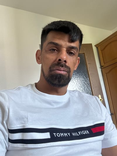 An adult male, appearing to be in his 20s or 30s, is depicted in a head-and-shoulders frontal shot. He has short, dark hair, dark eyes, and a neatly trimmed dark beard and mustache. His expression is neutral as he looks directly into the camera. He is wearing a white long-sleeved shirt or sweatshirt featuring a prominent horizontal black stripe across the chest, bordered by two thinner white stripes. The black stripe contains the embroidered white text "TOMMY HILFIGER" followed by a small red rectangular logo. The background on the left is a plain light-colored wall. On the right, a wooden door with a textured glass panel is partially visible behind his right shoulder, alongside a section of a light brown wooden cabinet or wardrobe. The indoor scene is located in Oberasbach, Germany.