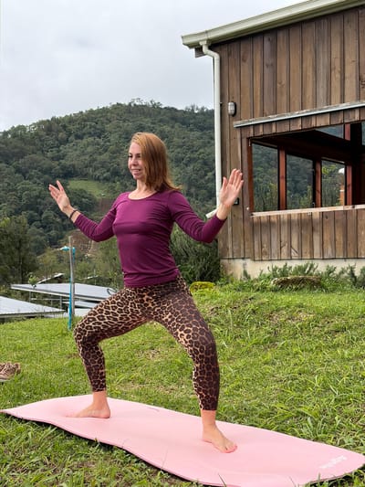 A woman with auburn hair stands on a pink yoga mat outdoors, performing a wide-legged squat with her arms raised. She is wearing a plum-colored long-sleeved shirt and leopard-print leggings. The background features a rustic wooden building with a window, and a lush, green hillside covered in trees and vegetation. The sky is overcast. In the distance, there appear to be some agricultural terraces. This scene is located in Ban Khun Wang, Thailand, a mountainous region known for its agricultural activities and cooler climate.
