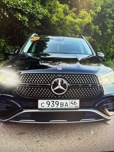 A black Mercedes-Benz GLE SUV is parked outdoors, with lush green trees in the background. The vehicle's front grille features the iconic Mercedes-Benz star emblem. A Russian license plate, displaying "C 939 BA 46" and the RUS flag, is prominently visible. A circular sticker with an orange and yellow design is affixed to the upper windshield. The overall lighting suggests it is daytime.