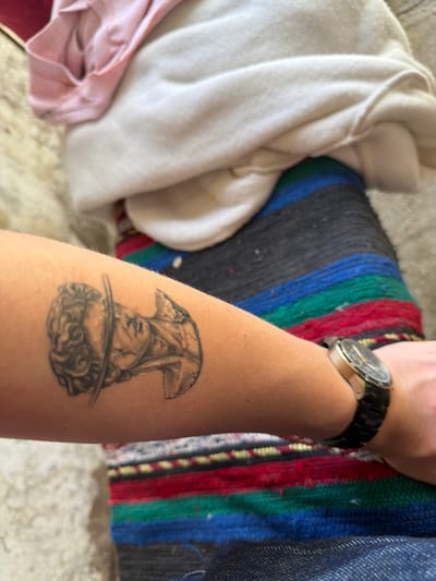 A close-up shot shows a forearm with a tattoo of an upside-down bust of a man, possibly a classical sculpture, positioned on the lower part of the arm. A gold-toned wristwatch with a black band encircles the wrist. Adjacent to the arm and tattoo are various items, including a light-colored, possibly white or cream-colored garment or fabric and a colorful striped blanket. The environment seems to be indoors. The image does not show the city of Kanchevo, Bulgaria, or any specific location. There are no people or actions depicted besides the presence of an arm with a tattoo.