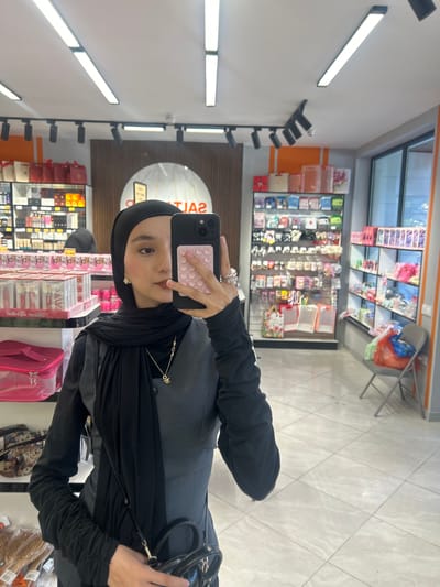 A person, wearing a black headscarf and a black dress, is taking a selfie in a retail establishment. The location is Ақсу, Kazakhstan. The subject is holding a mobile phone with a pink case. Aisles of retail products are visible in the background. The store has overhead fluorescent lighting. The floor is covered in tile. A black purse is visible on the right.