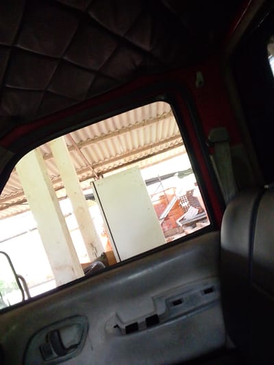 The image appears to be taken from inside a vehicle, looking out a window. The interior of the vehicle shows a padded headliner with a diamond pattern and a red frame around the window. The door panel is visible in the foreground, with a gray handle and some indentations. Outside the window, there are white pillars supporting a corrugated roof. Beyond the pillars, a brick structure with exposed rebar and scattered debris is visible. The overall environment suggests an outdoor, possibly semi-constructed or industrial setting, which could be found in a rural or semi-urban area like Atamo Sur, Venezuela. There are no people visible in the image.