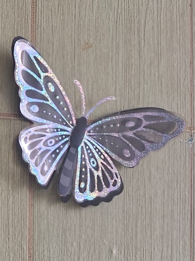 A holographic butterfly sticker is attached to a light-colored, vertically-paneled wooden surface. The butterfly is dark gray with iridescent wings featuring cut-out patterns and small, sparkling dots. The background appears to be a wall with a wood-grain texture. There are no people or animals present. The scene is indoors, possibly a room or a cabinet. The location in the city of Таёжное, Russia cannot be determined from the image.