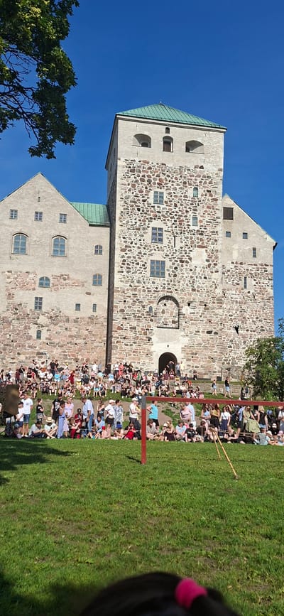 The image shows Turku Castle in Turku, Finland, under a clear blue sky on a sunny day. A large crowd of people are gathered on the grassy slope in front of the castle, many sitting or standing, watching an event. The castle itself is a large stone structure with a prominent tower and a green roof. The architecture suggests historical significance. The foreground shows a grassy area with some red and wooden poles that appear to be part of a barrier or stage setup. The overall scene depicts a public event or festival taking place at a historical landmark.