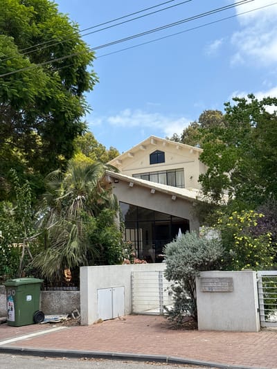 A house is surrounded by lush greenery and trees, under a sky with light clouds. The house features a distinctive triangular window and a large glass facade. A green trash bin sits near the sidewalk, while electrical wires stretch across the sky. The building facade has a textured finish.