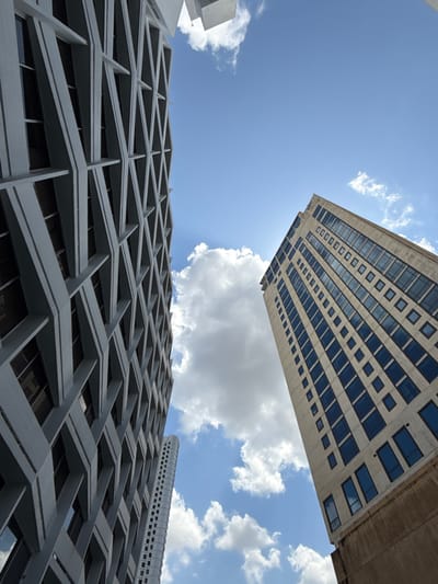 The image depicts an upward-looking view of three high-rise buildings against a bright blue sky with scattered white cumulus clouds, located in Bella Vista, Panama.
To the left, a tall, multi-story building features a light grey facade with a prominent geometric lattice structure composed of diagonal and horizontal concrete beams forming a grid pattern. Dark, recessed windows are visible behind this structure. This building extends out of frame at the top and bottom.
To the right, another tall building with a beige or light tan facade rises, characterized by numerous dark, rectangular windows arranged in vertical columns. The bottom visible section of this building transitions to a darker, possibly brown, base. This building also extends out of frame at the top and bottom.
Between these two primary structures, further in the background and towards the bottom, a slender, lighter-colored high-rise with many small, regularly spaced windows is partially visible. The sky is bright, with sunlight illuminating the clouds and parts of the buildings. No people or overt actions are visible in the scene.
