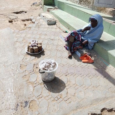 This image captures an outdoor scene in Dan Maliki, Nigeria. A person, likely a woman, with dark skin and dressed in a light blue head covering and a colorful patterned wrapper, is seated on the lowest step of a green-painted concrete staircase. She is barefoot, with her right arm raised towards her mouth, suggesting she is eating. A pair of orange open-toed sandals rests on the patterned concrete paving next to her.
On the ground in front of her, a white ceramic plate holds numerous stacks of round, light-brown food items, some dusted with white powder. Adjacent to the plate, a clear plastic bucket with a black handle contains a quantity of similar light-brown, irregular-shaped food items, also partially covered in white powder. The ground features worn, hexagonal concrete paving stones interspersed with areas of bare, dusty earth. The background reveals a building wall with a light-colored window frame and additional green steps. The scene is illuminated by strong sunlight, casting distinct shadows.