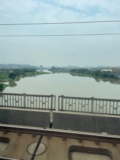 This is a media file depicting a wide, calm river flowing through a landscape with buildings and greenery on its banks. The perspective is from a bridge, evidenced by the concrete railing with a decorative pattern and a section of what appears to be a train car or similar structure on the right side.
The scene is somewhat hazy, suggesting either early morning, late afternoon, or a generally overcast and humid weather condition, typical of the region. The river reflects the sky and the structures along its edge. On the far bank, there are numerous buildings, ranging from low-rise structures to taller apartment complexes, indicating an urban or suburban area. Interspersed with the buildings are trees and patches of green land, possibly agricultural fields or parks. In the distance, rolling hills or mountains are faintly visible. The overall atmosphere is peaceful, with no obvious signs of human activity or movement, aside from the implied presence of the viewer on the bridge.
The time of day is not precisely determinable due to the haze but appears to be during daylight hours. The location cues such as the architectural style of the buildings and the presence of the river suggest an East Asian setting, consistent with the provided context of Dongguan, China. There is no discernible text within the image. The railing of the bridge, the reflective surface of the water, and the distant cityscape are the most prominent visual elements.