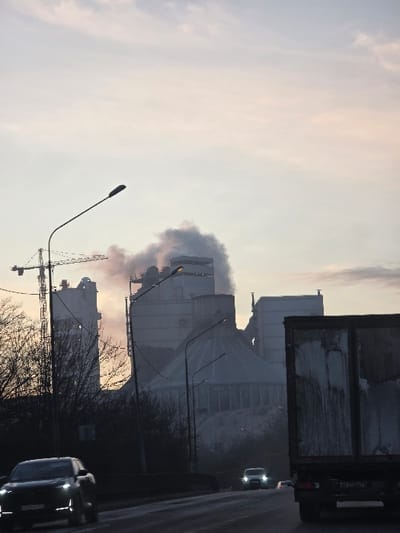 The image presents a wide view of an industrial landscape in Verkhnebakanskiy, Russia, under overcast skies at what appears to be dawn or dusk. The sky exhibits pale blue, pink, and white tones. In the foreground, a two-lane asphalt road runs horizontally, appearing wet or reflective. A dark-colored SUV with illuminated LED headlights is observed in motion on the left lane, driving towards the viewer. Further down the road, another vehicle with illuminated headlights is visible in the distance. On the far right, the rear section of a large, dark truck, possibly a semi-trailer, is visible in the right lane, facing away from the viewer; its cargo area shows dirt and residue. Two streetlights with modern fixtures are positioned on the left, arching over the road. Bare deciduous trees and shrubs line the left side of the road. The background is dominated by a large industrial facility, identified as a cement plant. It comprises numerous tall, grey concrete silos, processing towers, and a prominent conical structure. A large tower crane is visible on the far left of the complex. Multiple plumes of thick, white-grey smoke or steam are actively emitted from various points within the plant, rising against the dim sky, indicating ongoing operations. Several structures within the plant are artificially illuminated. Large piles of material are discernible around the base of the industrial structures. No people are visible.