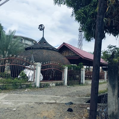 The image captures a static outdoor scene featuring a mosque complex in Kuta Pungwa, Indonesia, under a cloudy sky. The primary subject is a mosque building with a distinctive, large, dark-tiled, dome-like roof, possibly made of shingles or scales, crowned with a weathered metal crescent moon and star finial. A decorative fringe runs along the base of this dome.
In front of the mosque is a rusty metal fence with pointed decorative elements, set between concrete posts. Behind this fence, some greenery and shrubs are visible. To the right of the main dome, another building with a steep, red-tiled gabled roof is partially seen, featuring intricate wooden carvings under its eaves. Further back and to the right, a tall, slender metal lattice minaret extends into the sky.
On the left side, lush green palm fronds and other tropical tree leaves obscure part of another rounded building with multiple arched windows adorned with white, patterned grilles. A thick, dark utility pole stands prominently on the right side of the foreground, partially obscuring the view and rising towards other power lines crossing the top left of the frame.
The ground in the foreground consists of a rough dirt and gravel path or road, with patches of concrete and scattered debris like straw and dark fabric. No human activity or visible text is present in the image. The diffused lighting suggests an overcast daytime setting.