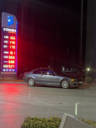 The image displays a nighttime scene at a Struma Petrol gas station located in Piperkov Chiflik, Bulgaria. On the left, a tall, illuminated gas price totem prominently displays "STRUMA PETROL" with a blue "S" logo at the top. Below, red LED digits indicate various fuel prices: 100 octane at 1.66, 95 octane at 1.46, Diesel Plus at 1.70, Diesel at 1.61, LPG at 0.66, and AdBlue at 0.98. A green icon with "0.37" is visible at the bottom of the price list.

A dark gray two-door coupe, identifiable as a BMW E46 3-Series, is parked stationary on the paved forecourt, angled slightly toward the camera and facing right. The vehicle features multi-spoke alloy wheels, and a Bulgarian license plate, 'CTA 7404 BP', is visible on its front bumper. The background is dark, with faint lights and indistinct structural outlines suggesting an urban environment beyond the gas station. Low green bushes are partially visible and out of focus in the immediate foreground. No individuals are present or engaged in any activities within the frame.