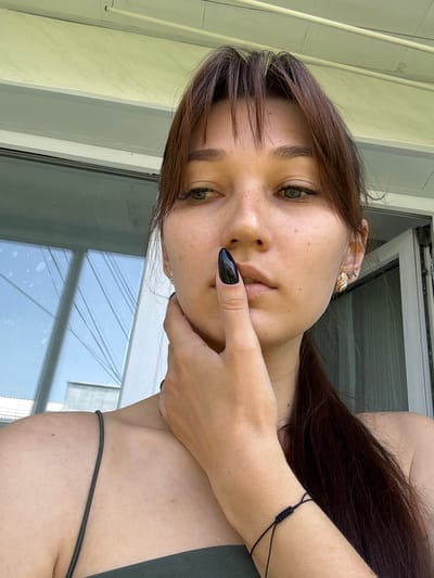A young woman with fair skin and brown hair, pulled back into a ponytail, is captured in a close-up, outdoor portrait. She has light green eyes and a smattering of freckles across her nose and cheeks. Her left hand is raised, with a long, black, almond-shaped nail on her thumb touching her lips, while her left hand supports her chin. She is wearing a dark green, strappy top and a gold earring with a diamond-shaped design in her right ear. A black string bracelet is visible on her left wrist. The background suggests a residential setting, possibly a balcony or porch, with a window reflecting a light blue sky with some white streaks, and part of a white building. The overall lighting indicates daytime. The image does not provide any specific visual cues to pinpoint its location within Novokuznetsk, Russia.