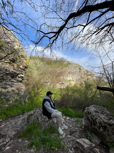 A man is sitting on a rock outcrop in Barkach, Bulgaria. He is wearing a dark baseball cap, a gray hooded sweatshirt, a black vest, tan pants, and white sneakers. He is sitting on a gray, rocky surface covered with green vegetation. Behind him is a steep, rocky cliff face covered with green trees and blooming white flowers. The sky is visible above the cliff face through the branches of bare trees.