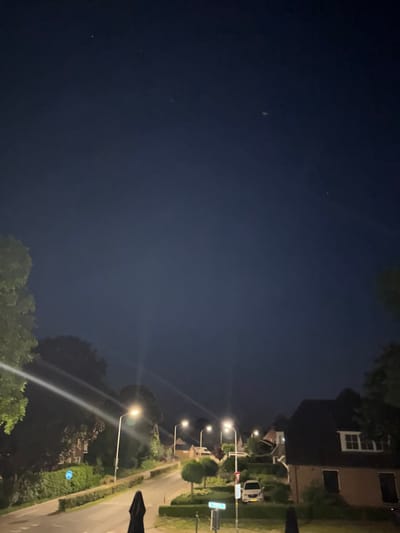 The image depicts a nighttime street scene in the city of Silvolde, Netherlands. The sky is dark blue with a few visible stars. The street is illuminated by streetlights, casting beams of light. Several houses with lit windows are visible on either side of the street, and some trees and manicured hedges line the roadside. Two cars are parked on the street, and a bicycle path sign is visible on the left side of the road. A dark, draped object, possibly a parasol, is situated on the right side of the foreground.