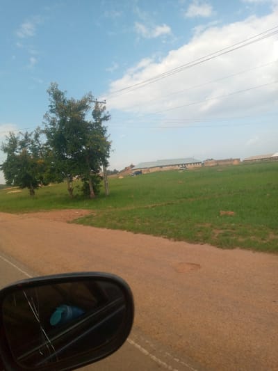 The image shows a view from a car looking towards a grassy field with several buildings in the distance. Two trees are prominent in the foreground on the left side of the frame. A side mirror of the car is visible in the lower left corner, showing a cracked reflection of a blue object. The road in front of the car is brown and appears to be paved. In the background, there are low-lying buildings with light-colored roofs, and utility wires are visible strung across the sky. The sky is blue with scattered white clouds. The scene appears to be in a rural or semi-rural setting, possibly on the outskirts of a city like Rahwol Chom, Nigeria, given the presence of buildings and utility infrastructure.