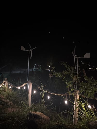 At night in Khlong Khian, Thailand, the scene features vegetation along a water's edge, illuminated by string lights. Two small, white wind turbines are visible along the shoreline. The background is dark, suggesting a rural or minimally lit location. The plants include tall grasses, foliage and what appears to be bamboo poles.