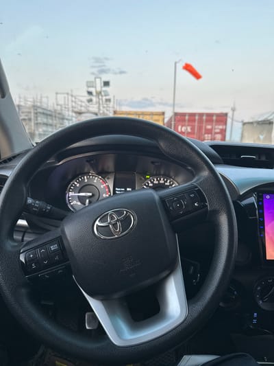 The image, taken from inside a Toyota vehicle, shows a view through the windshield. The dashboard is visible, featuring a steering wheel with the Toyota logo, instrument cluster, and various control buttons. Outside the vehicle, a construction site is apparent with scaffolding and cranes. A bright orange windsock is visible against the sky, indicating wind. Shipping containers, one with the "tex" logo, are also present. The sky is overcast with clouds. The location is identified as Қосшағыл ауылдық округі, Kazakhstan.