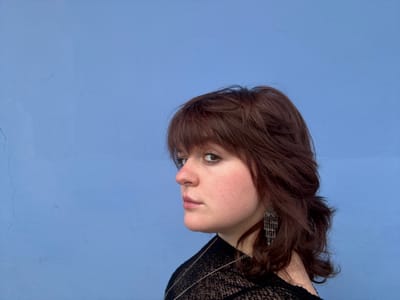 A portrait of a woman with auburn, shoulder-length hair is shown against a light blue background. She has fair skin, freckles, and is wearing an earring. A black, lacey top is visible. Her gaze is directed to the right of the frame. The image is oriented upside down.