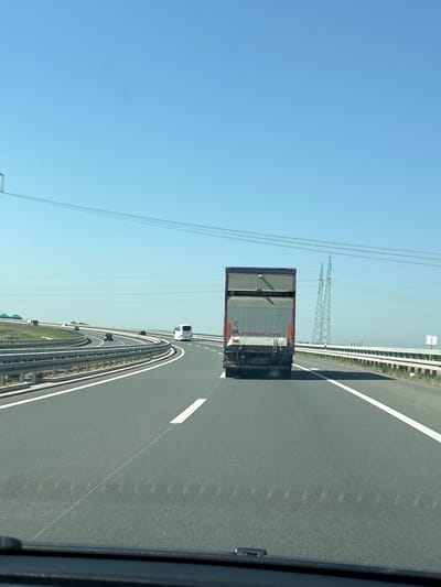 The image depicts a wide, multi-lane highway under a clear blue sky. The perspective is from inside a vehicle, looking forward. A large, silver truck with a dark-colored trailer is prominent in the center of the frame, traveling in the same direction as the viewer. The truck has some markings on its rear, including "LEVende dyr" and a circular sign with a number inside.

To the right of the truck, a tall, lattice-like electrical pylon stands in the distance, with power lines stretching across the horizon. A white signpost with "km 3" is visible along the right side of the highway. The highway itself has clear white lane markings and is bordered by guardrails and a white line. In the distance, on the left side of the highway, several other vehicles, including a white bus and smaller cars, are visible, navigating a curve. The environment appears to be a rural or suburban area, with flat terrain and some greenery visible beyond the highway.

While the image shows a highway setting, there is no specific visual information within the image to definitively place the location in Ruma, Serbia. The "km 3" sign might suggest a distance marker, but without further context or recognizable landmarks specific to Ruma, this cannot be confirmed.