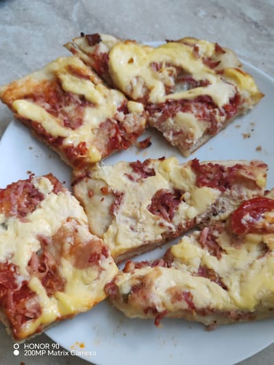 The image shows several slices of pizza arranged on a white plate. The pizza has a crust, tomato sauce, a generous amount of melted cheese, and visible pieces of meat, likely ham or bacon, and possibly some tomato slices. The pizza appears to be homemade or from a casual eatery, with uneven crust edges and toppings spread across the surface. The background is a textured surface, possibly a countertop or table, with a light brown and gray marble pattern. There is no visible indication of the city of Krasnaya Polyana, Russia, or any people in the image. The bottom left corner of the image contains a watermark that reads "HONOR 90 200MP Matrix Camera".