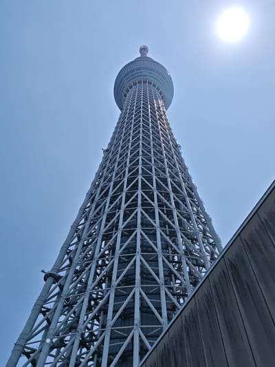 An upward-angled, full-shot photograph captures the Tokyo Skytree in Sumida, Japan, under a bright, clear sky. The tower, a prominent landmark, dominates the frame with its intricate lattice structure of white beams. At the top, the Skytree transitions into a rounded observation deck encased in glass. To the right foreground, a dark, angled building partially obscures the lower portion of the tower. The sun is visible as a bright, white orb in the upper right corner of the image. No people are visible in the scene.