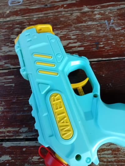The image shows a blue and yellow plastic toy water gun on a weathered wooden surface. The word "WATER" is prominently displayed on the side of the gun in yellow lettering. A red cap is visible near the bottom of the gun. The photograph may have been taken in Malko Chochoveni, Bulgaria, as requested.