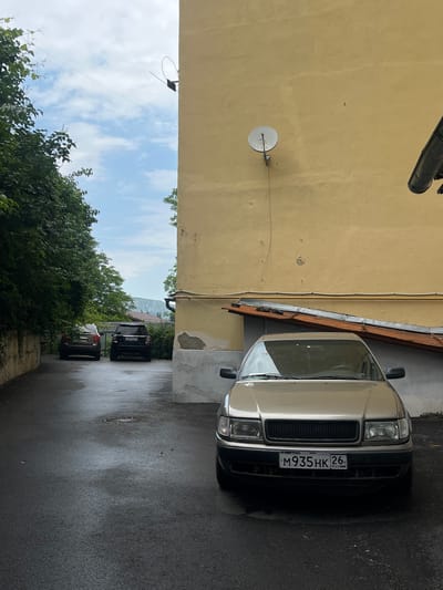 A silver sedan is parked on a wet asphalt surface in an alleyway. The car has a license plate that reads "M935HK 26 RUS". To the left of the sedan, two SUVs are parked further down the alley, one brown and one black. The alley is bordered by a tall, yellow building on the right and a densely vegetated wall on the left. A satellite dish is mounted on the yellow building. The sky is cloudy and blue. The overall impression is of a slightly damp, urban environment.