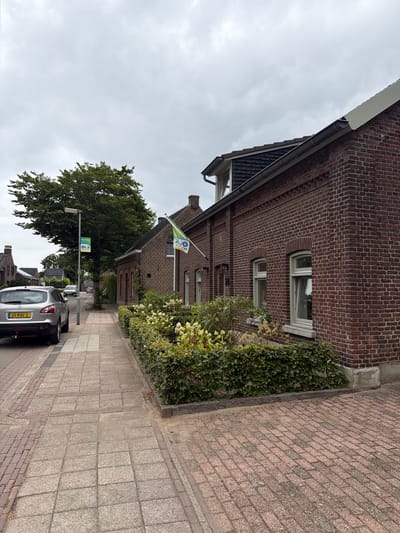The image depicts a street scene in Melick, Netherlands, on an overcast day. In the foreground, a silver Nissan Qashqai with license plate 33-RXL-2 is parked on the left side of the street. To the right of the car, a paved sidewalk runs along a brick building. A street lamp with a sign that reads "OLS MELICK" is positioned next to the sidewalk.
The building on the right is constructed of red brick and features multiple windows with light green frames. A green and white flag is flying from a flagpole attached to the building. In front of the building, a well-maintained hedge with flowering plants forms a border along the sidewalk.
The street is paved with reddish-brown bricks, and the sidewalk is composed of grey concrete pavers with some brick detailing. In the background, more houses and trees can be seen, and the sky is heavily clouded. The scene appears to be a quiet residential street.