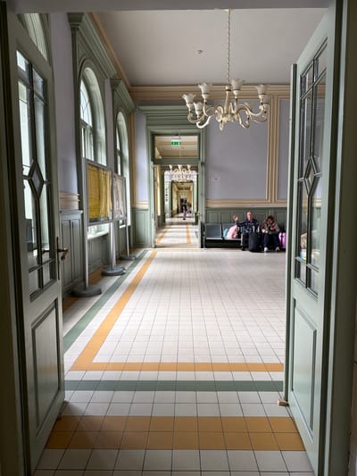 The image shows a long, narrow corridor with white tiled floors with alternating stripes of yellow and green. The walls are light grey and feature arched windows and doorways. Doors are open on both sides of the corridor. In the far distance, people are seated on chairs in a waiting area. A chandelier hangs from the ceiling in the foreground. The image appears to be taken from inside a room looking down the corridor. The location is Przemyśl, Poland.