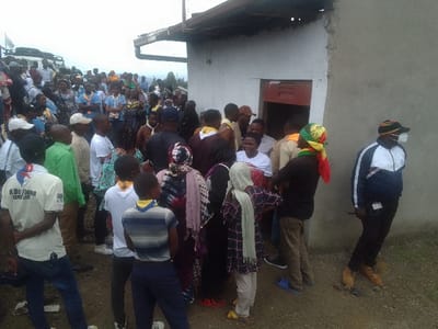 A crowd of people, many wearing blue and yellow scarves, gather near a white building with a red door. Some individuals are dressed in casual clothes, while others wear scout uniforms. A man with a colorful headwrap stands near the building's corner. The atmosphere is casual. The building appears to be a small structure, and the crowd seems to be waiting or congregating for an event.