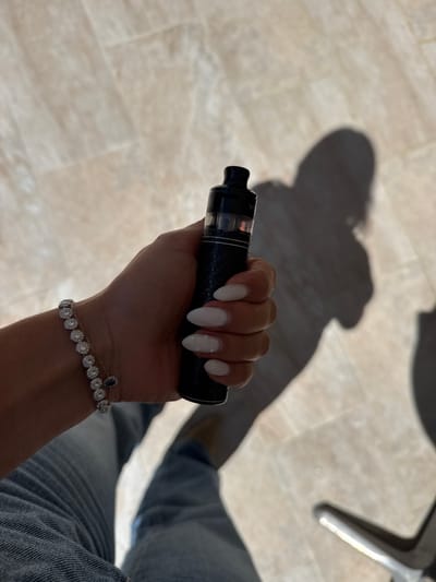 A top-down shot shows a person's hand holding a vape pen. The hand has almond-shaped white nails, one with a scattering of white dots. A bracelet with small, circular, diamond-like stones and a single teardrop-shaped blue stone adorns the wrist. The person is wearing light blue denim jeans. The background is a beige tiled floor with a chair leg visible in the upper right corner. The image has strong shadows.