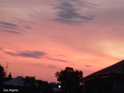 A photograph taken in Kuje, Nigeria captures a sunset over a residential area. The sky is dominated by hues of pink and orange, with streaks of gray clouds present. In the foreground, silhouetted against the vibrant sky, are the dark outlines of trees and rooftops. One prominent rooftop is angled towards the right side of the frame, revealing a dark roof with visible structural lines. The text "Gui, Nigeria" is superimposed in white text in the lower left corner of the image. The overall scene depicts a tranquil evening in a Nigerian town.