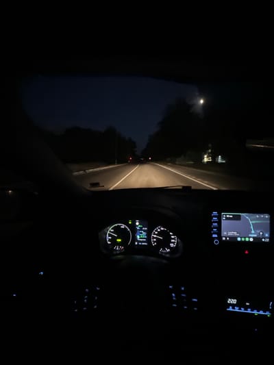 This image captures a view from inside a car at night, looking forward along a dimly lit road. The interior of the car is dark, with illuminated displays and controls providing the main light sources. A navigation screen displays a map with a route and estimated travel time. The instrument cluster features a speedometer showing a speed of 60 km/h and a digital display indicating 22.0 degrees Celsius. Other buttons and controls are also illuminated in the darkness. Outside the car, the road is flanked by dark trees, with a faint light visible in the distance. The scene suggests the car is in motion on a highway or a country road. The time appears to be late evening or night.