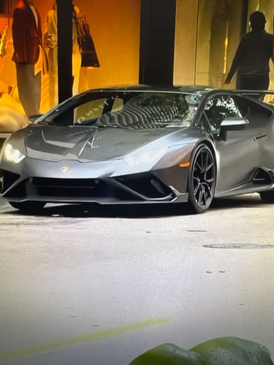 The image depicts a dark gray sports car, likely a Lamborghini, parked outdoors. The car's sharp, aerodynamic design is visible, with a prominent front bumper and a detailed wheel. Reflections on the car's surface suggest it is daytime, and the lighting implies clear weather. To the left of the car, there is a blurred background of a building interior, possibly a store or showroom, with golden and dark horizontal stripes. A silhouetted figure is partially visible within this interior space, standing near the car. On the right side of the image, a large, out-of-focus green object, possibly a plant or piece of produce, is present. The setting appears to be urban, with a textured pavement surface where the car is parked. The overall impression is a snapshot of a high-performance vehicle in an everyday setting, possibly in Chorzów, Poland. No visible text is discernible in the image.