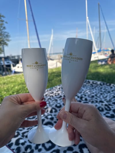 This media file shows two white champagne flutes with the "MOËT & CHANDON CHAMPAGNE" logo printed in gold. The flutes are held by two hands, one visible with red nail polish. The background is blurred but suggests an outdoor setting with green grass and blue sky with some blurry masts of sailboats, indicating a sunny day. The image is likely taken in Gstadt am Chiemsee, Germany, given the context of sailboats and a lakeside setting. The scene conveys a sense of celebration or a relaxed, upscale outdoor gathering.