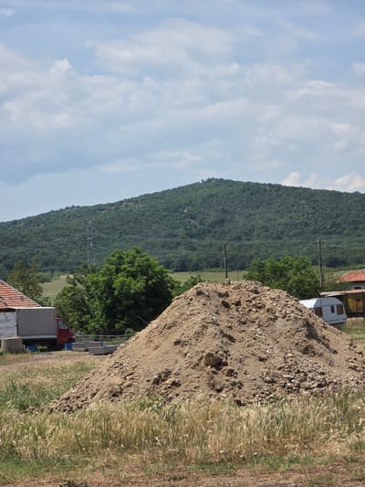 The image captures a rural landscape in Ivan Vazovo, Bulgaria, dominated by natural elements and some signs of human activity. The scene is presented from a slightly elevated perspective, looking towards a forested mountain range under a partly cloudy sky.
In the foreground, a large, light brown mound of earth or gravel occupies the right side of the frame. To its left, a dense growth of tall, dry, straw-colored grass and weeds stretches across the midground, bordering a narrower strip of greener vegetation.
The midground features several elements indicating human presence. Behind the foreground mound, a white caravan or trailer is partially visible, along with parts of a reddish-brown tiled roof of a building and a red truck. Green trees with full foliage are scattered throughout this area, one prominently positioned towards the left of the dirt mound. Multiple utility poles and power lines crisscross the midground horizontally, suggesting a power infrastructure.
In the background, a large, dark green mountain, densely covered in trees, rises prominently and extends across the left side of the image. The upper portion of the frame is filled with a bright blue sky, adorned with scattered white and wispy clouds.
The photograph depicts a bright daytime scene, likely under clear weather conditions. No specific human activity is observable, and no visible text is present. The overall impression is one of a natural environment coexisting with some rural development or construction.
