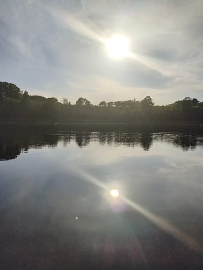 This image depicts a serene lake scene in Brocēni, Latvia, likely during late afternoon or early evening given the low angle of the sun and the warm, diffused light. The sky is partly cloudy, with the bright sun creating a prominent lens flare that extends across the upper portion of the frame. The sun's reflection is also visible on the water's surface, creating a shimmering effect and another lens flare in the lower half of the image.
The foreground is dominated by the dark, reflective water, which mirrors the sky and the tree line. A dense line of trees and foliage forms the background, separating the lake from the sky. A few small structures or buildings appear to be nestled within the trees on the distant shore. There are no distinct people or objects clearly visible in the main body of the lake, though there may be some very small, indistinct shapes on the water surface suggesting distant boats or buoys. The overall mood of the image is peaceful and tranquil, emphasizing the natural beauty of the landscape. There is no visible text in the image.