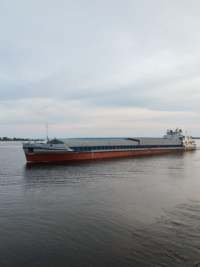 A long cargo ship, possibly a river barge, moves through a body of water, likely a river or a large lake. The ship features a grey front section with the Cyrillic word "СИММАХ" visible, followed by a long, flat cargo area covered in grey panels and what appears to be blue containers. The hull is painted red. The rear of the ship is white with an upper deck. The water is dark, with ripples indicating movement. The sky is overcast with grey and white clouds. The location is Суда, Russia, as the ship is labeled with the name in Cyrillic. The surrounding land is barely visible in the distance, suggesting a wide body of water. The scene appears to be captured during daylight.